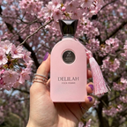 Frasco do perfume Delilah da Maison Alhambra em ambiente externo com flores cor-de-rosa.